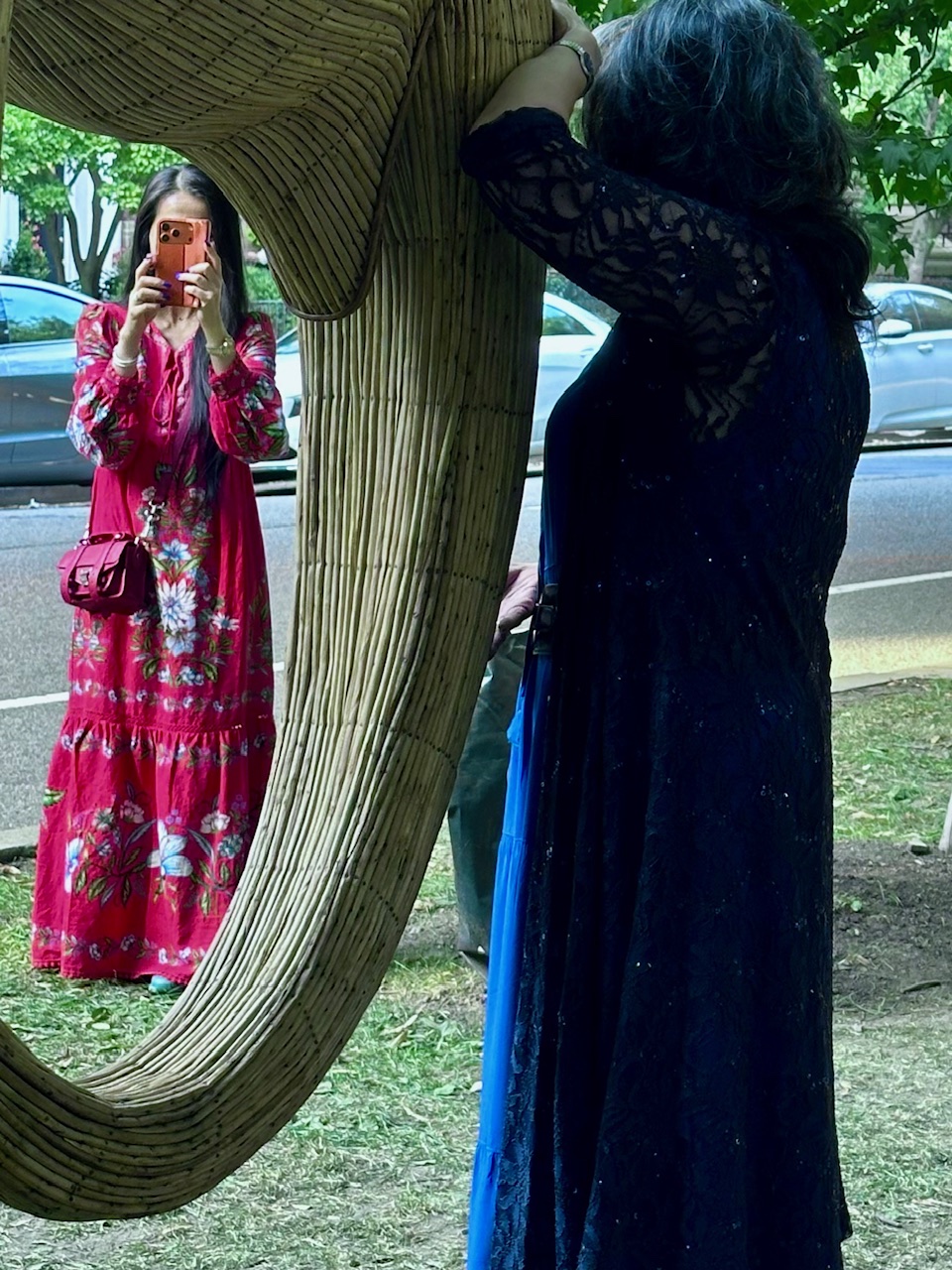 Two visitors on the Commonwealth Avenue Mall standing beside an elephant sculpture's raised trunk, one photographing the other through the curve.