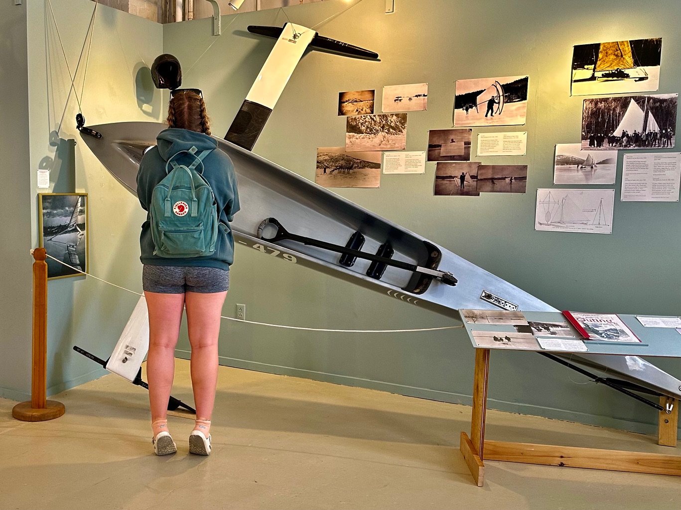A visitor with a backpack studying a slender ice boat mounted on the gallery wall in the Chilly Chronicles exhibit, with historic winter photographs displayed behind it.
