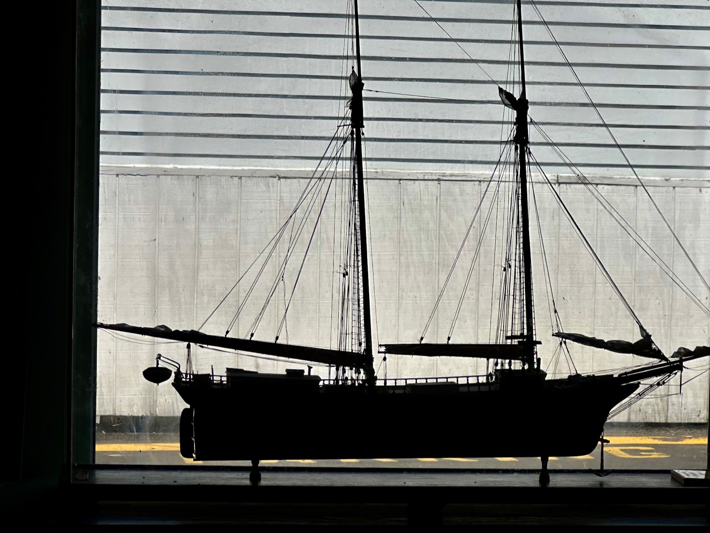 A rigged model schooner silhouetted in a museum window, its masts and standing rigging dark against the bright wharf outside.