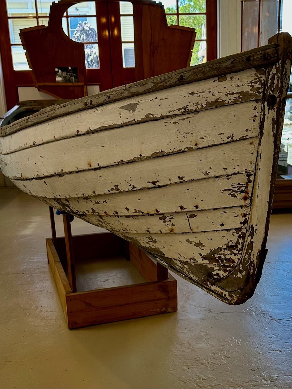 The weathered white planked bow of a wooden skiff resting on a cradle in the museum gallery, its paint flaking back to bare wood along the seams.