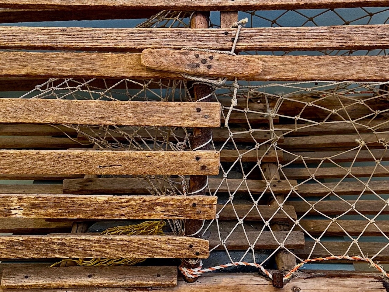 Close view of a wooden lobster trap, its oak laths, knotted netting, and orange and white twine filling the frame.
