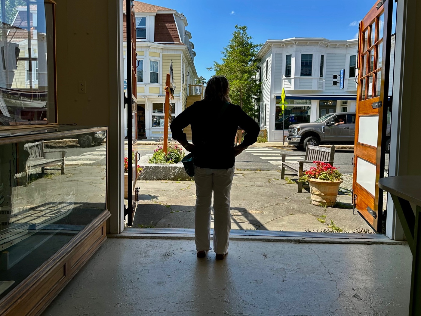 A woman standing in the museum's open doorway with her hands on her hips, looking out at the clapboard storefronts and crosswalk of Main Street in Northeast Harbor.