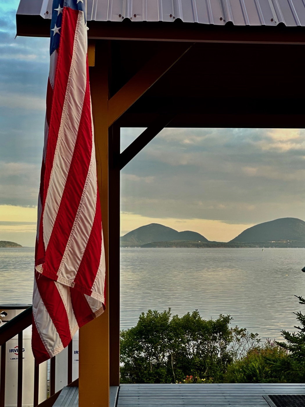An American flag hanging from a porch post in late afternoon light, framing the rounded hills of islands across the water Downeast.