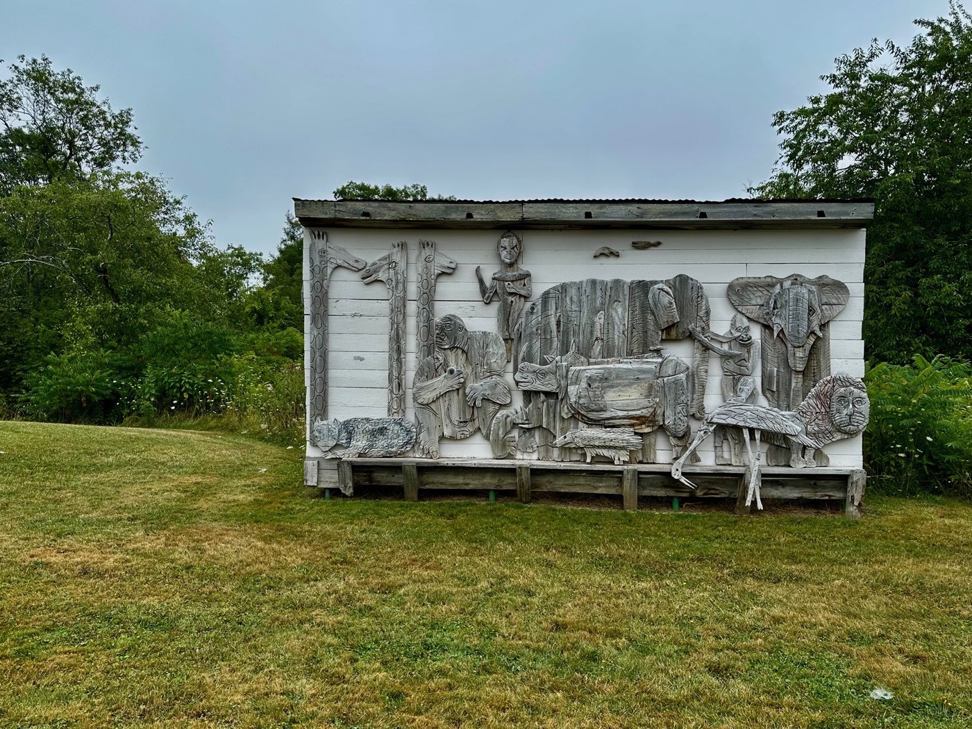 A long white barn wall covered end to end with carved wood relief animals in weathered gray, among them an elephant, a lion, giraffes, a rhinoceros, and a monkey, set above a mowed field.