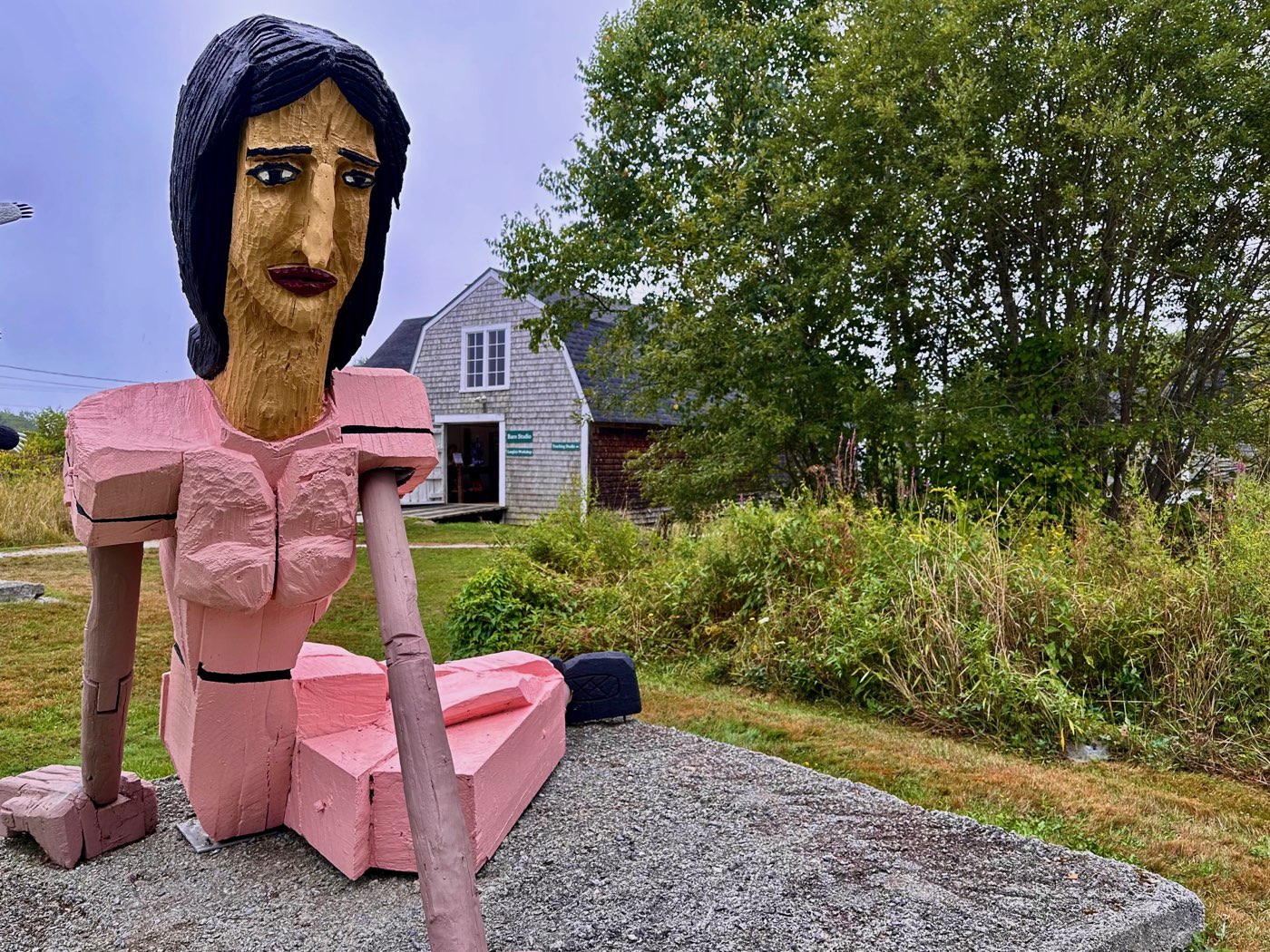 A large carved wood figure of a seated woman painted pink, with black hair and a staff across her lap, resting on a gravel mound in front of the shingled studio barn.