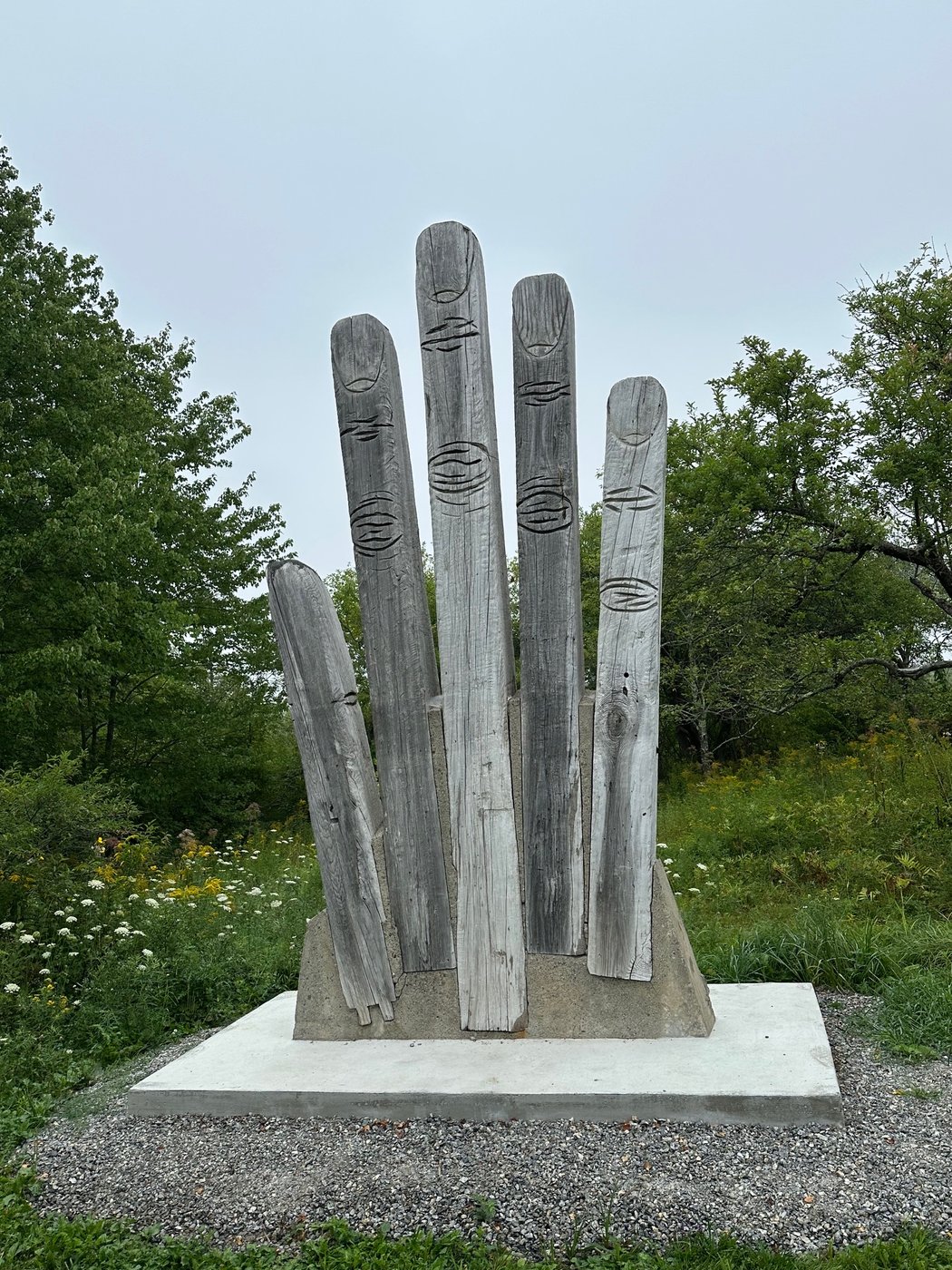 Five weathered gray wood planks set upright on a concrete plinth, each carved with simple eye shapes, standing against a background of trees and wildflowers.