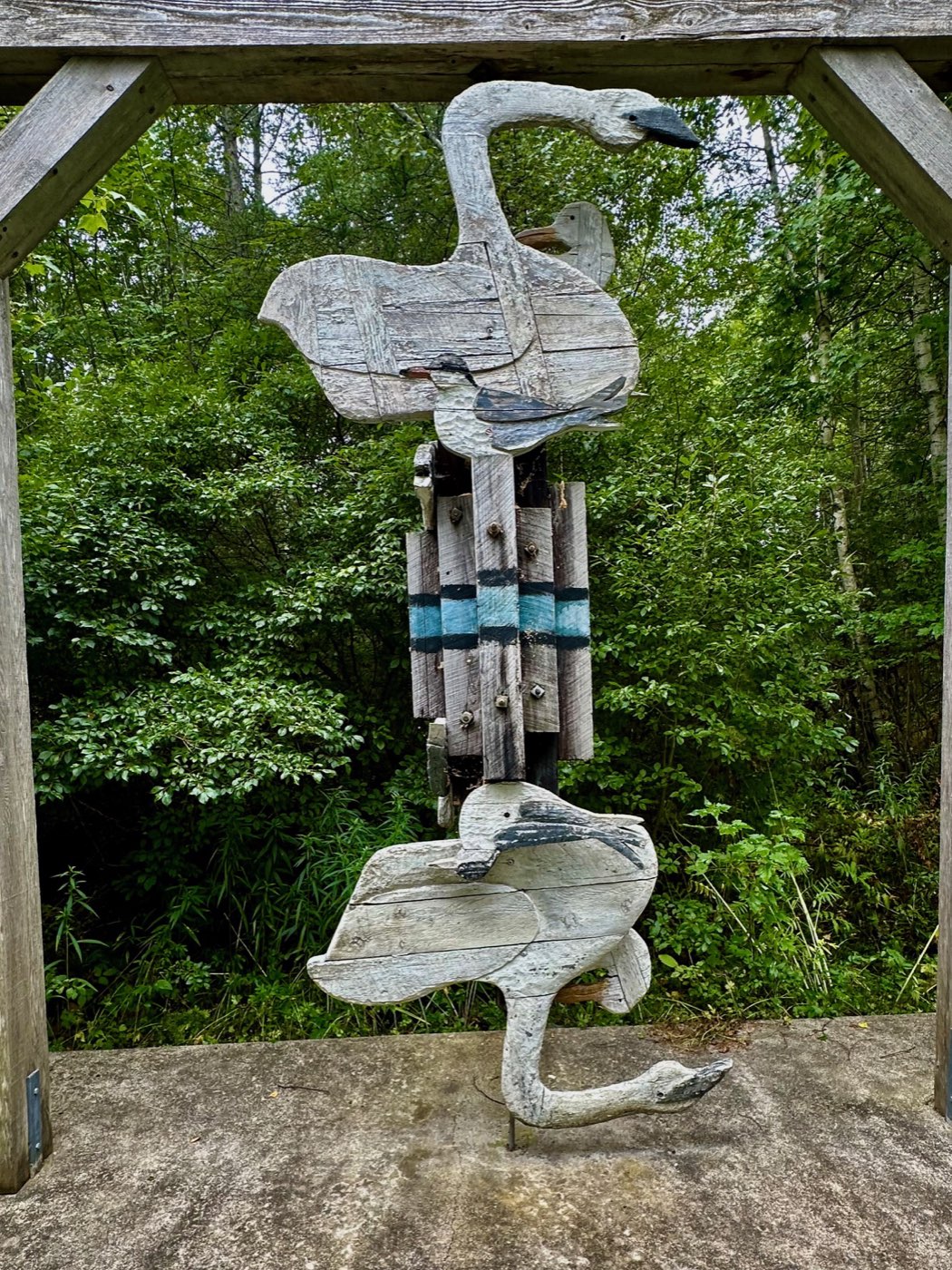 Carved wood swans and shorebirds mounted on a weathered post, seen through the timber frame of a viewing shelter with dense green woods behind.