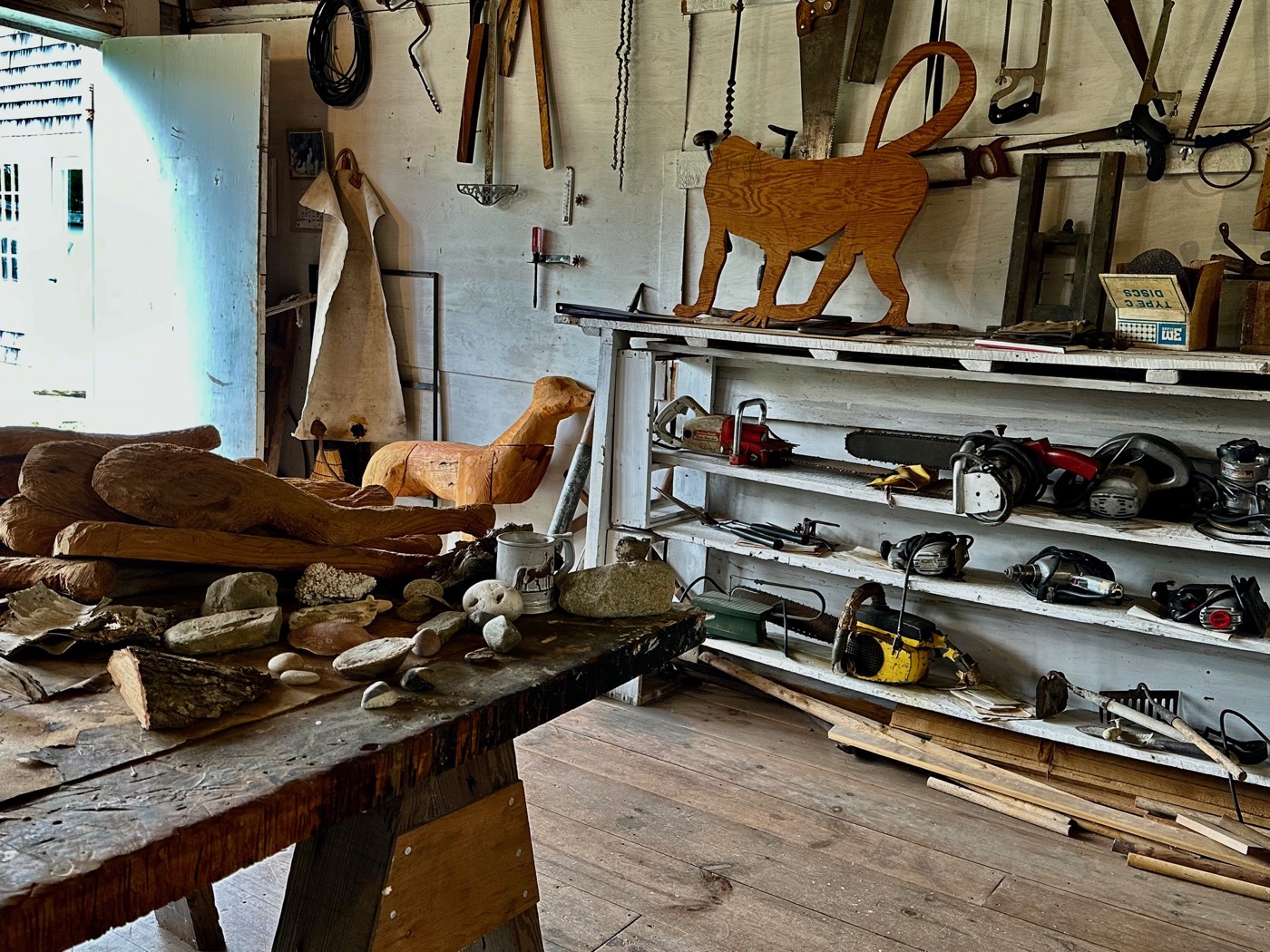 The interior of Bernard Langlais&rsquo;s studio, hand saws and chainsaws racked on white shelves, a carved wood cat cutout above them, and roughed out wood forms and beach stones covering the workbench.