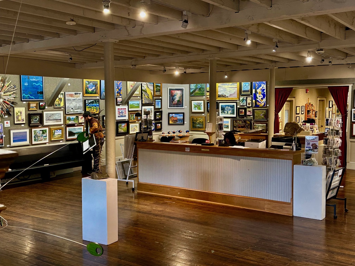The beamed main gallery of the North Shore Arts Association in Gloucester, with salon-hung walls and a reception desk.