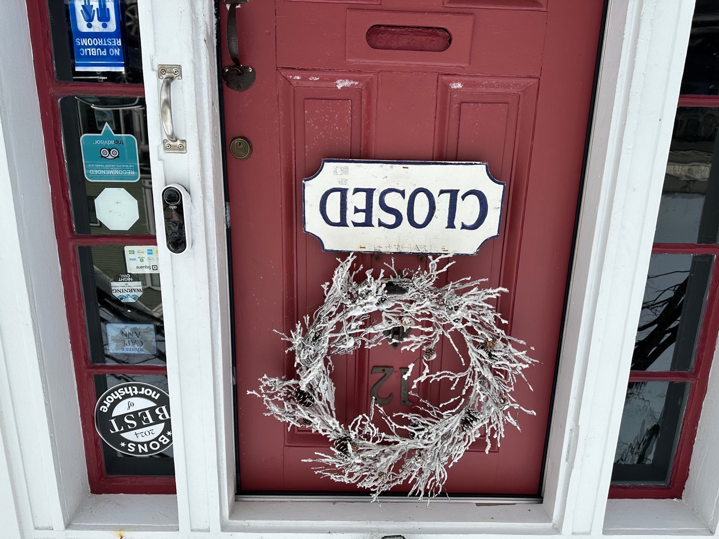 An upside-down Closed sign and a winter wreath on the weathered red door of the Rockport Art Association.